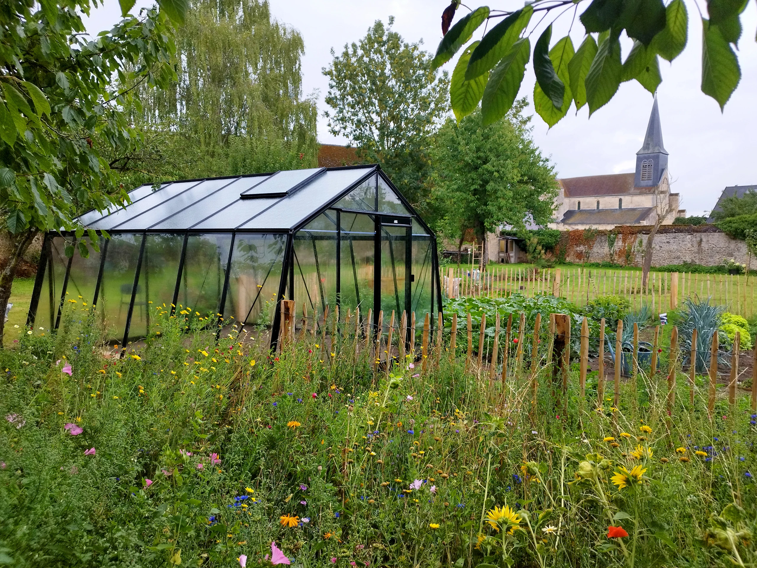 Serre de jardin en aluminium noir installée dans un potager fleuri avec vue sur clocher de village à Ham — Colver