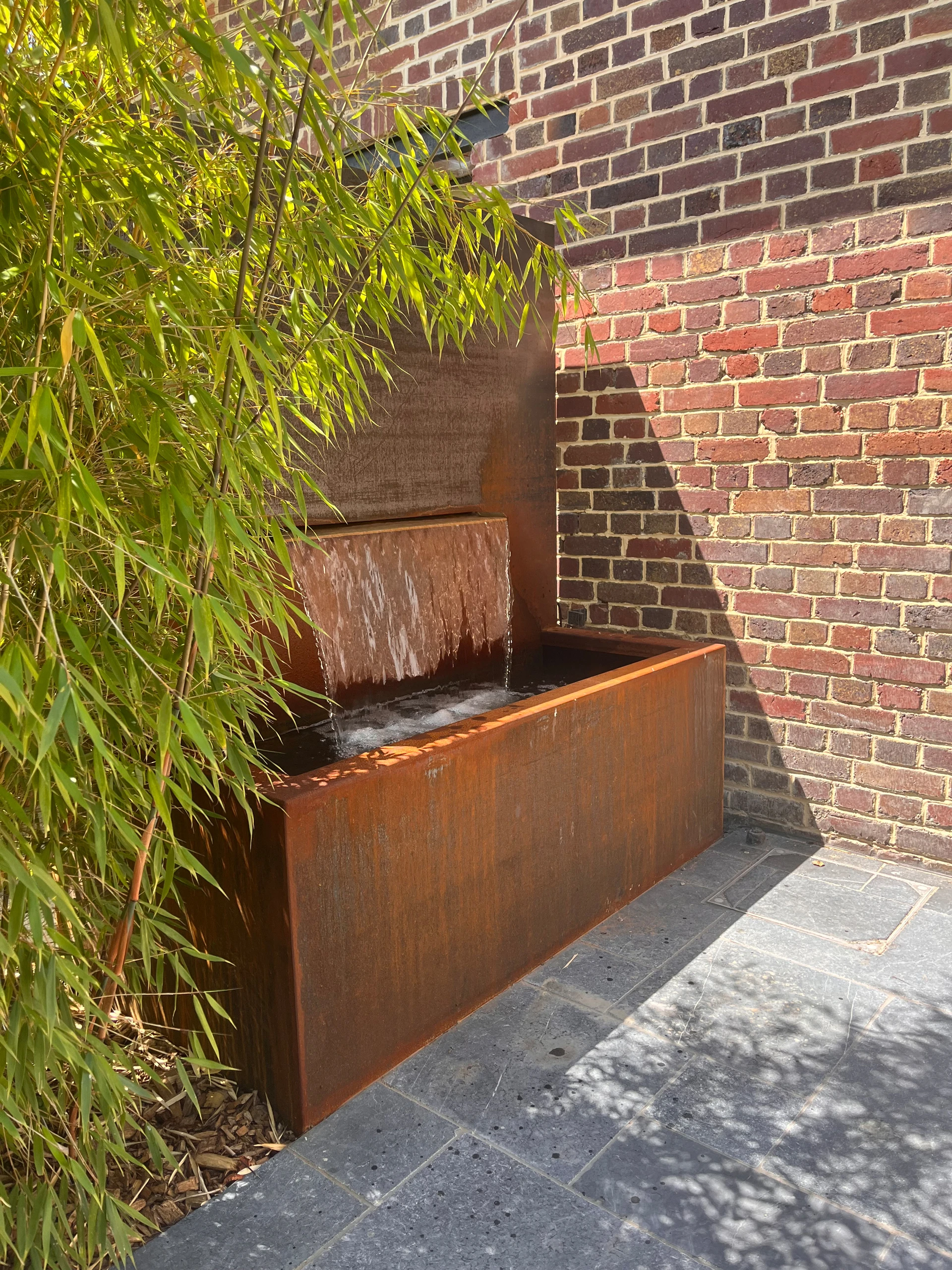 Fontaine lame d'eau en acier Corten en cascade sur terrasse ardoise, contre mur de briques avec bambou — Roye, Somme — Colver